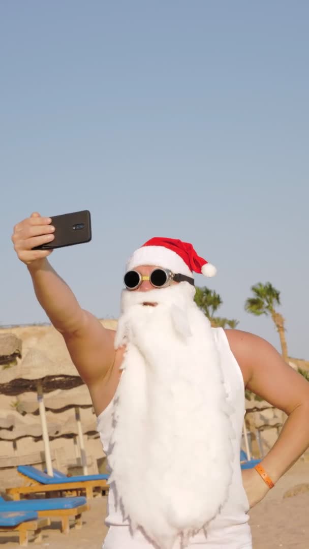 Santa Claus takes selfie. vertical video. Santa summer vacation. Santa Claus blogger. Funny Santa, in sunglasses, takes selfie video on beach by the sea, against palm trees backdrop. — Stock Video