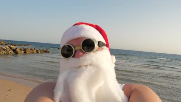 Blogerka Świętego Mikołaja. Święty Mikołaj robi selfie. Śmieszny Mikołaj, w okularach przeciwsłonecznych, kręci selfie na plaży nad morzem, na tle palm. Santa letnie wakacje. — Wideo stockowe