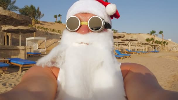 Weihnachtsmann-Blogger. Der Weihnachtsmann macht Selfie. Der lustige Weihnachtsmann mit Sonnenbrille macht Selfie-Videos am Strand am Meer vor Palmen-Kulisse. Weihnachtsmann-Sommerurlaub. — Stockvideo