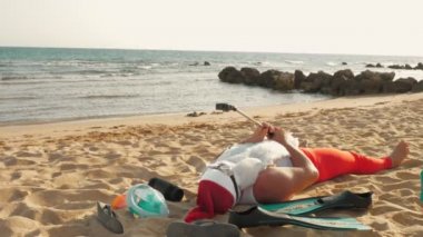 Noel Baba blogcusu. Noel Baba selfie çeker. Güneş gözlüklü komik Noel Baba, deniz kenarındaki kumlu sahilde uzanırken selfie videosu çekiyor. Noel Baba yaz tatili.