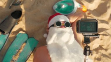 Noel Baba blogcusu. Noel Baba selfie çeker. Güneş gözlüklü komik Noel Baba, deniz kenarındaki kumlu sahilde uzanırken selfie videosu çekiyor. Üst Manzara. Noel Baba yaz tatili.