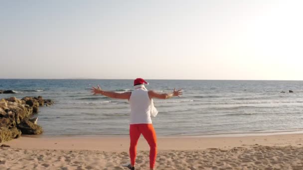 Wakacje Świętego Mikołaja. Mikołaj dobrze się bawi. Zabawny Mikołaj, w płetwach biegnie przez piaszczystą plażę do morza. Spieszy mu się do pływania, nurku. widok z tyłu — Wideo stockowe