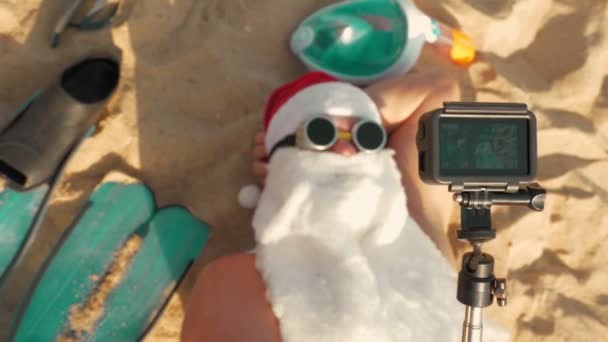 Blogerka Świętego Mikołaja. Święty Mikołaj robi selfie. Śmieszny Mikołaj, w okularach przeciwsłonecznych, robi selfie wideo, leżąc na piaszczystej plaży nad morzem. Widok z góry. Santa letnie wakacje. — Wideo stockowe