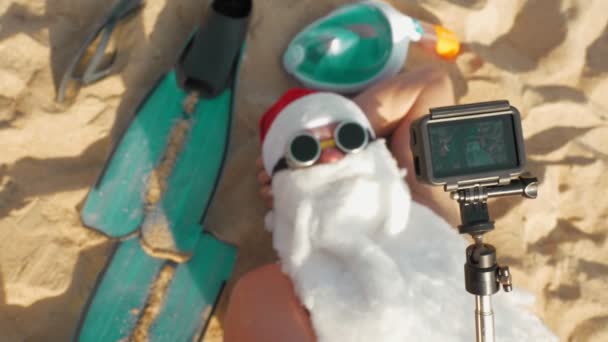Blogueur du Père Noël. Le Père Noël prend du selfie. Santa drôle, dans les lunettes de soleil, prend selfie vidéo, tout en étant couché sur la plage de sable près de la mer. vue de dessus. Vacances d'été Santa. — Video