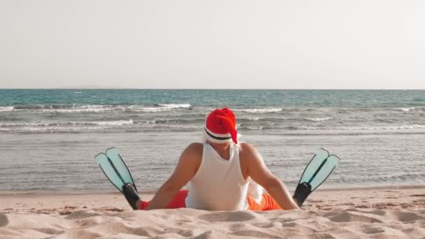 Wakacje Świętego Mikołaja. Święty Mikołaj dobrze się bawi. Śmieszny Mikołaj, w płetwach, relaksujący siedząc na piaszczystej plaży nad morzem. widok z tyłu. — Wideo stockowe