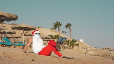 Noel Baba yaz tatili. Noel Baba eğleniyor. Koruyucu maskeli, güneş gözlüklü ve paletli komik Noel Baba deniz kenarında palmiye ağaçlarının arasında uzanırken dinleniyor.