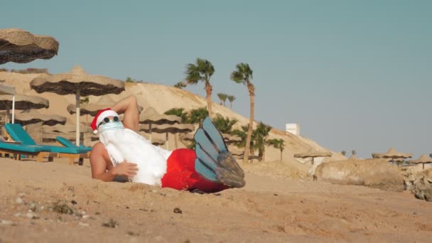 Moş Crăciun vacanţă de vară. Moş Crăciun se distrează. Moș Crăciun amuzant, în mască de protecție, ochelari de soare și papuci, relaxant în timp ce se află pe plajă de nisip lângă mare, printre palmieri — Videoclip de stoc