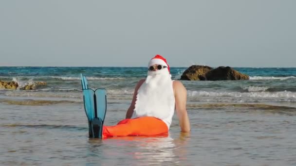 O Pai Natal engraçado. Pai Natal, em óculos de sol e nadadeiras, sentado e se divertindo, relaxando na água do mar perto da praia de areia. Papai Noel férias de verão — Vídeo de Stock