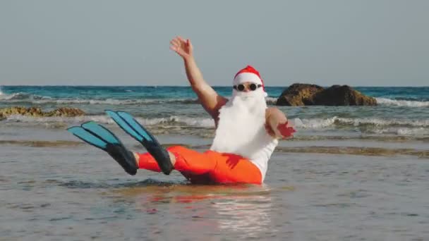 O Pai Natal engraçado. Pai Natal, em óculos de sol e nadadeiras, sentado e se divertindo, relaxando na água do mar perto da praia de areia. Papai Noel férias de verão — Vídeo de Stock