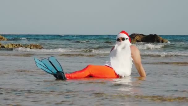 O Pai Natal engraçado. Pai Natal, em óculos de sol e nadadeiras, sentado e se divertindo, relaxando na água do mar perto da praia de areia. Papai Noel férias de verão — Vídeo de Stock