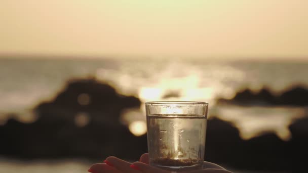 Vaso de agua al atardecer. Primer plano. silueta de una mano femenina sostiene un vaso con agua clara. fondo de una puesta de sol o salida del sol sobre el mar — Vídeos de Stock