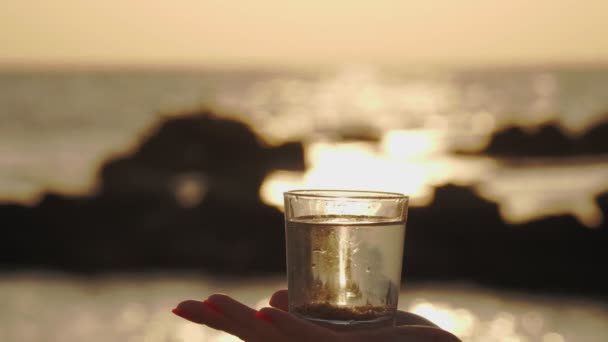Bicchiere d'acqua al tramonto. primo piano. la sagoma di una mano femminile tiene un bicchiere con acqua limpida. sfondo di un tramonto o alba sul mare — Video Stock