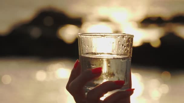 Vaso de agua al atardecer. Primer plano. silueta de una mano femenina sostiene un vaso con agua clara. fondo de una puesta de sol o salida del sol sobre el mar — Vídeos de Stock