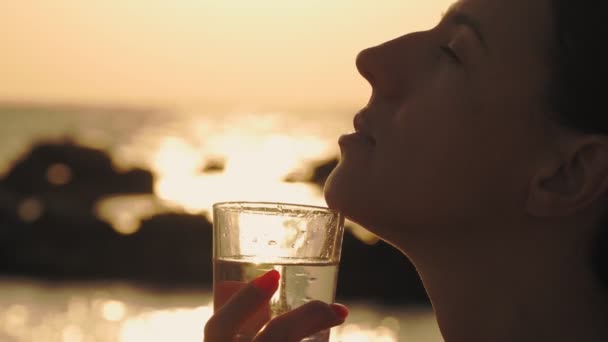 Bicchiere d'acqua al tramonto. primo piano. silhouette. donna tiene un bicchiere di acqua limpida vicino al viso di profilo. sfondo di un tramonto o alba sul mare — Video Stock