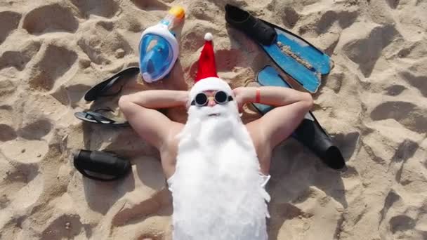 Ο Άγιος Βασίλης, φορώντας γυαλιά ηλίου, βρίσκεται στην αμμώδη παραλία δίπλα στη θάλασσα και κάνει ηλιοθεραπεία. Στην κορυφή. Καλοκαιρινές διακοπές. — Αρχείο Βίντεο