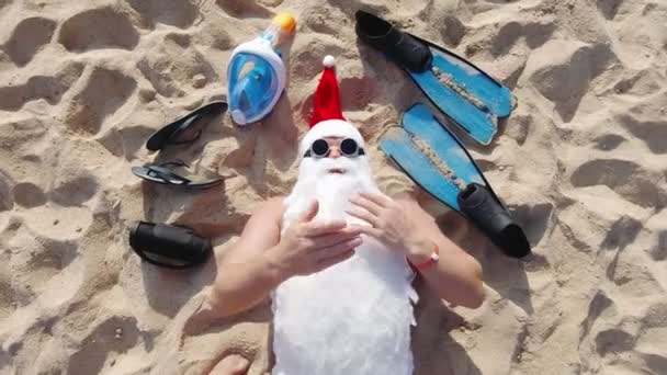 Санта Клаус, у сонцезахисних окулярах, лежить на піщаному пляжі біля моря і б'є поцілунками, щоб відтворити відеокамеру. вид зверху. Санта-Літня відпустка. Блог Санта Клауса.. — стокове відео