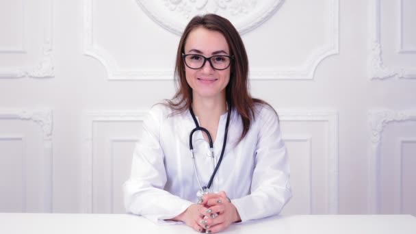 Conseil et thérapie en ligne. Jeune femme médecin en uniforme médical blanc avec stéthoscope parlant par vidéoconférence à l'aide d'un ordinateur, assise devant webcam, dans une clinique de santé ou à l'hôpital — Video