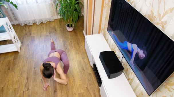 Online fitness edzés távolról, otthon. Fiatal nő, sportruházatban, oktatóvideót néz fitneszedzővel a TV-ben és otthon gyakorolja a gyakorlatokat. Online edzés. — Stock videók