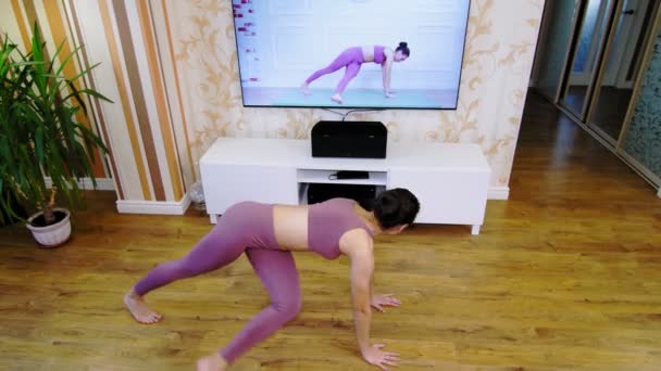 Allenamento fitness online da remoto, a casa. Giovane donna, in abbigliamento sportivo, guardando video tutorial con allenatore di fitness in TV e praticando esercizi a casa. Allenamento online. — Video Stock