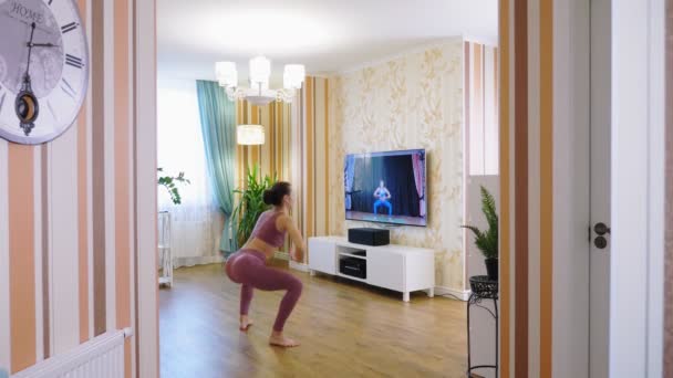 Entrenamiento de fitness en línea de forma remota, en casa. Mujer joven, en ropa deportiva, viendo video tutorial con entrenador de fitness en la televisión y practicando ejercicios en casa. Entrenamiento en línea. — Vídeo de stock