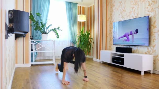 Online προπόνηση, εξ αποστάσεως. Νεαρή γυναίκα, στα αθλητικά, βλέποντας βίντεο με φροντιστήριο γυμναστικής στην τηλεόραση και εξάσκηση ασκήσεων στο σπίτι. κάνοντας αθλητισμό κατά τη διάρκεια του αποκλεισμού, στο σπίτι — Αρχείο Βίντεο