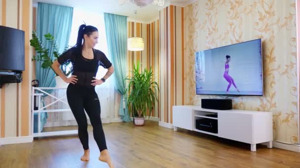Online workout, remotely. Young woman, in sportswear, watching tutorial video with fitness coach on TV and practicing exercises at home. doing sports during lockdown, at home — Stock Video