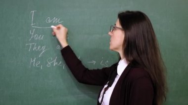 Çevrimiçi eğitim. Bayan öğretmen çevrimiçi İngilizce dersi veriyor. Okulda, tahtaya tebeşirle dil bilgisi kuralları yazıyor. Uzaktan eğitim. Sınıftan canlı video yayını.