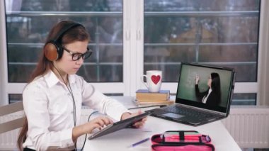 Uzaktan öğrenme, online eğitim. Öğrenci kız, kulaklıkla dizüstü bilgisayarda video dersi izliyor ve tabletine notlar alıyor. Evde online ders çalışıyorum..