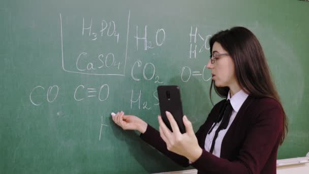 Online onderwijs. Docent houdt online scheikundeles voor studenten, op afstand, met behulp van smartphone. Ze schrijft formules met krijt op schoolbord, in de klas. onderwijs op afstand. live video — Stockvideo