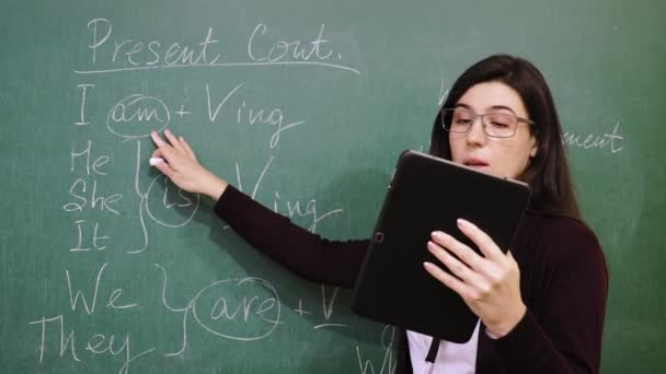 Educación en línea. El profesor está impartiendo clases de inglés en línea para estudiantes, de forma remota, utilizando una tableta digital. Ella está escribiendo reglas gramaticales con tiza en la pizarra, en la clase escolar. enseñanza remota. en vivo — Vídeo de stock