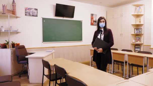 Enseignement à distance, confinement dans les écoles. femme professeur, dans un masque de protection, se tient dans une salle de classe vide de l'école avec un ordinateur portable dans ses mains. verrouillage complet dû au coronavirus. — Video