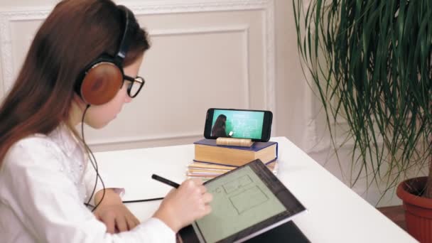 Afstandsonderwijs, online onderwijs. schoolmeisje, in koptelefoon, kijken naar online tutorial video-les op de smartphone en het maken van notities op haar tablet. thuis online studeren. — Stockvideo