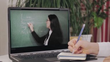 Uzaktan öğrenme, online eğitim. Laptop ekranına yakın çekim. Öğrenci kız öğretmenlerin açıklamaları ve not defterine not düşmesiyle çevrimiçi video dersleri izliyor. Evde online ders çalışıyorum..