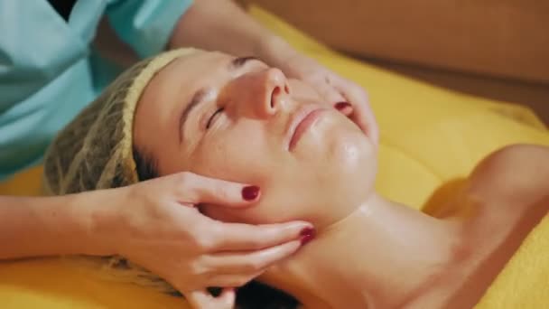 Zabieg odmładzania, masaż twarzy. zbliżenie. kobieta leży na kanapie i masaż twarzy do tonizowania mięśni i drenażu limfatycznego skóry twarzy, w klinice kosmetycznej. Pielęgnacja skóry, kosmetologia — Wideo stockowe