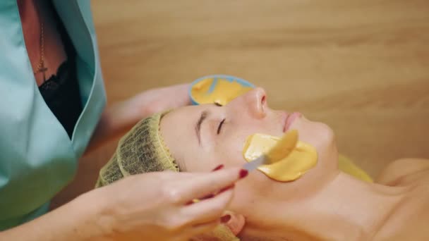 Kosmetologi, prosedur perawatan kulit wajah. close-up. ahli kecantikan menerapkan emas alginate topeng kosmetik untuk wajah wanita. Kosmetik anti-penuaan. — Stok Video