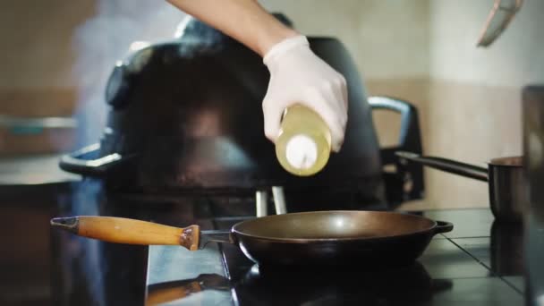 Cookery. the cook, chef, in protective gloves, pours cooking oil, vegetable oil from bottle into a hot frying pan. frying food. — Stock Video
