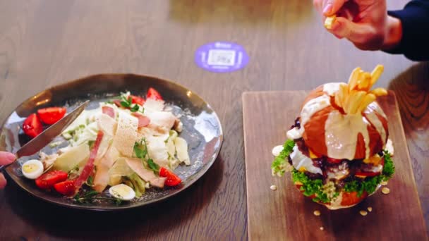 Salat und Burger. Nahaufnahme. Im Café stehen ein Teller Salat und ein großer saftiger Burger mit Soße auf dem Tisch. Die Menschen essen zu Mittag. Gesundes Essen versus Fast Food, Junk Food. — Stockvideo