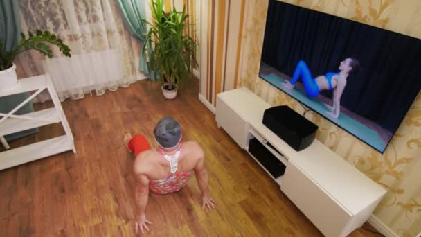 Funny freak. Fat man in multicolored T-shirt and red tight-fitting leggings, is engaged in fitness, fooling around at home. cheerful guy entertains himself, doing aerobic exercises to lose weight, on — Stock Video