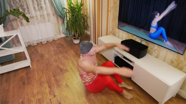 Funny freak. Fat man in multicolored T-shirt and red tight-fitting leggings, is engaged in fitness, fooling around at home. cheerful guy entertains himself, doing aerobic exercises to lose weight, on — Stock Video