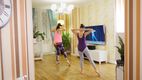 Fitnesz otthon. Fiatal nő és tizenéves lány, sportkosztümökben, oktatóvideót néz fitneszedzővel a TV-ben és otthon gyakorolja a gyakorlatokat. Online edzés, távolról. — Stock videók