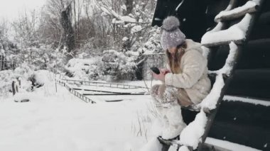 Karlı kış eğlencesi. Sıcak kış kıyafetleri içinde, kar taneleriyle kaplı sevimli bir kız, bankta oturuyor ve akıllı telefon kullanıyor, açık havada, kar yağarken, eski ahşap evin yakınında. karlı kış