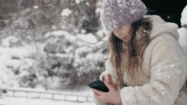 Kış portresi. Kışlık kalın giysiler içinde, kar taneleriyle kaplı sevimli küçük bir kız, bankta oturuyor ve akıllı telefon kullanıyor, açık havada, kar yağarken, eski ahşap evin yakınında. karlı