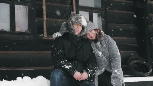 Kış portresi. Sıcak kışlık giysiler giymiş, kar taneleriyle kaplı mutlu, birbirine aşık bir çift, eski bir ahşap evin yanında oturuyorlar ve sarılıyorlar. Karlı havada mutlu zamanlar — Stok video