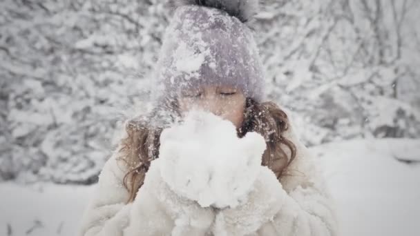 雪の冬の楽しみ。雪が吹いた。暖かい冬の服を着て、カメラ、屋外、雪の公園や森の中で手から雪を吹いています。. — ストック動画