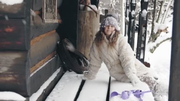 Retrato de inverno. gracinha, menina bonita, vestida com roupas quentes de inverno, totalmente coberta com flocos de neve, sentada em um banco por uma velha casa de madeira. tempo feliz no dia de inverno nevado. — Vídeo de Stock