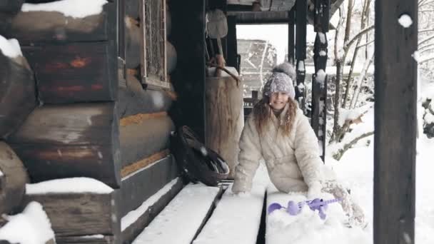 Kış portresi. Şirin, güzel, kışlık giysiler içinde, kar taneleriyle kaplı, eski ahşap bir evin yanındaki bankta oturan bir kız. Karlı kış gününde mutlu günler.. — Stok video