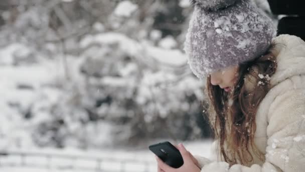 Kış portresi. Kışlık kalın giysiler içinde, kar taneleriyle kaplı sevimli küçük bir kız, bankta oturuyor ve akıllı telefon kullanıyor, açık havada, kar yağarken, eski ahşap evin yakınında. karlı — Stok video