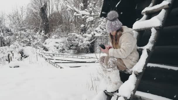 Śnieżna zimowa zabawa. Cutie girl, ubrana w ciepłe zimowe ubrania, w pełni pokryta płatkami śniegu, siedzi na ławce i za pomocą smartfona, na zewnątrz, podczas opadów śniegu, w pobliżu starego drewnianego domu. śnieżna zima — Wideo stockowe