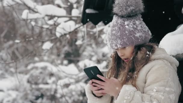 Kış portresi. Kışlık kalın giysiler içinde, kar taneleriyle kaplı sevimli küçük bir kız, bankta oturuyor ve akıllı telefon kullanıyor, açık havada, kar yağarken, eski ahşap evin yakınında. karlı — Stok video