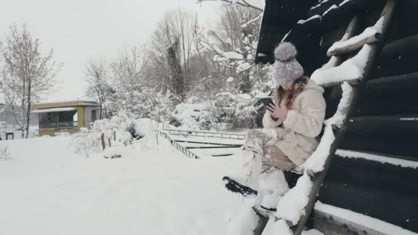 Śnieżna zimowa zabawa. Cutie girl, ubrana w ciepłe zimowe ubrania, w pełni pokryta płatkami śniegu, siedzi na ławce i za pomocą smartfona, na zewnątrz, podczas opadów śniegu, w pobliżu starego drewnianego domu. śnieżna zima — Wideo stockowe